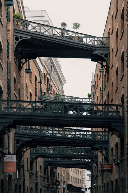 A series of black metal bridges connecting two adjacent brick buildings in Bermondsey, London, forming a layered walkway above a narrow street. The bridges feature decorative railings and are adorned with potted plants, with natural light illuminating the scene. The image captures the architectural detail and urban environment, reflecting the typical setting for residential or commercial buildings in the area, as cleaned and maintained by Carpet Cleaners Bermondsey during their surface cleaning and sanitisation services.