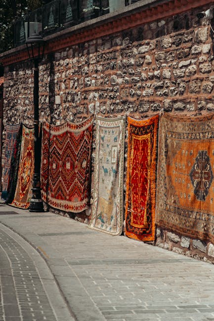 A row of colorful, intricately patterned rugs hanging along a rustic stone wall on a paved sidewalk, with natural daylight illuminating the scene. The rugs vary in design, featuring geometric and floral motifs in shades of red, orange, blue, and beige. The setting appears to be an outdoor marketplace or street stall, with the rugs displayed for sale or display purposes. The stone wall behind them adds a textured, historic feel. Carpet Cleaners Bermondsey is mentioned as the service provider, emphasizing domestic cleaning and surface maintenance in the context of the page about Bermondsey Street carpet cleaning prices and offers.