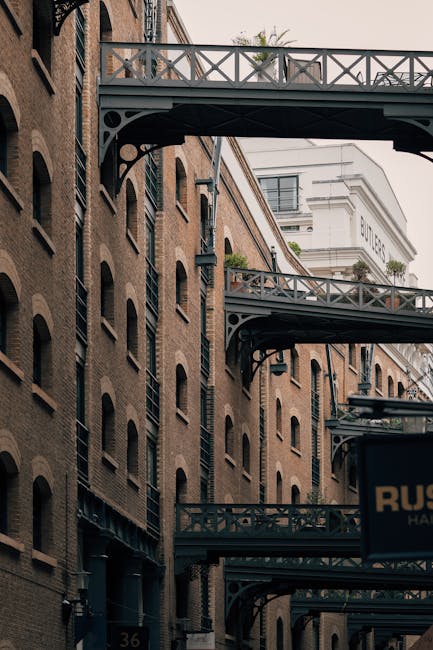An empty cobblestone street in Bermondsey, featuring brick buildings on either side with black metal fire escape bridges spanning between them. The scene is illuminated by natural daylight, highlighti